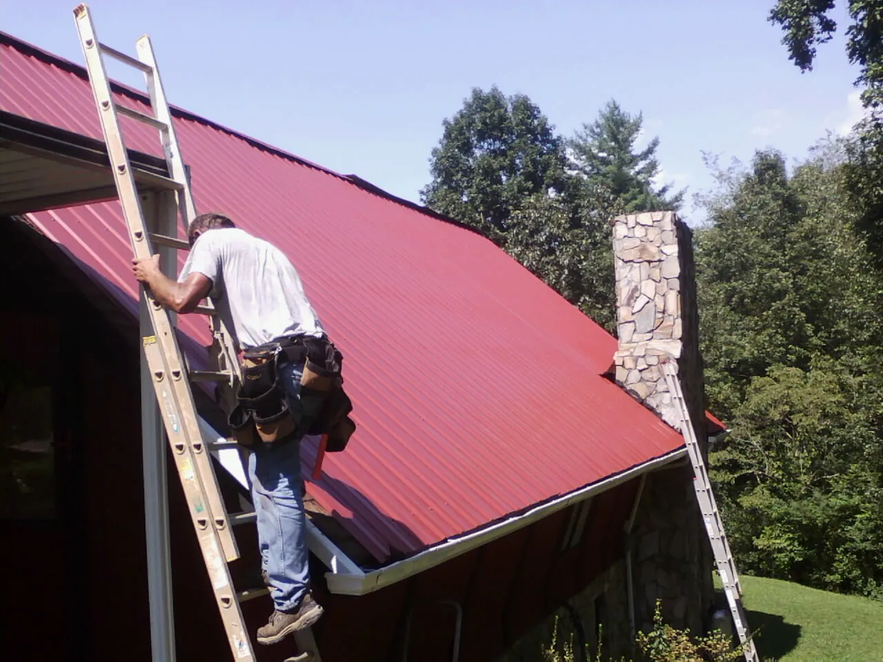 Roofer on a ladder beside a red metal roof and stone chimney — Soffit & Fascia Repair by Roofing Brothers in Bunnlevel